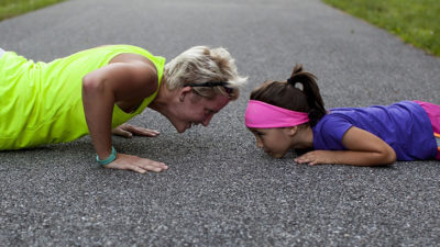 En vuxen person och ett barn ligger mitt emot varandra på marken. De gör push ups och har träningskläder.