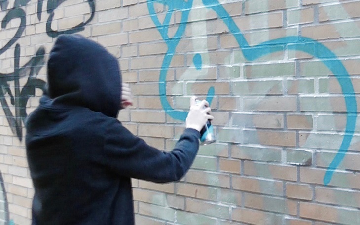 En person med mörk tröja och luvan uppdragen står vänd mot en vägg. Vi ser inte ansiktet. Personen har en sprejburk i handen och sprejar på en tegelvägg.