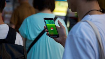 Ryggar på flera människor. Den som står närmast har en mobil i handen. På skärmens syns Pokémon Go-spelet.