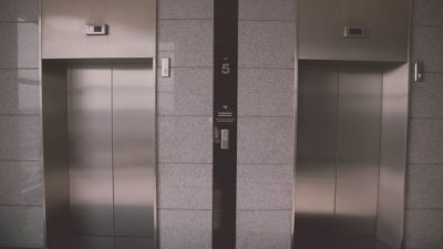 Bilden föreställer två hissdörrar, sida vid sida. Dörrarna är gjorda i ett silvrigt metallmaterial. Runt om dem är det grått kakel.