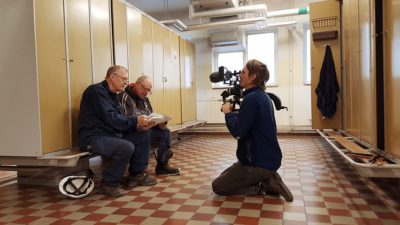 Ett omklädningsrum. På en bänk framför en rad med skåp sitter två äldre män. De har blå overaller på sig och tittar på ett foto. Framför dem sitter Nils Petter Löfstedt på knä och filmar dem med sin kamera.