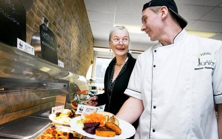 Två vuxna personer tittar på varandra och ler. De står framför en lunchservering. Den ena håller upp en tallrik med mat. Den andra lägger upp mat på sin tallrik.