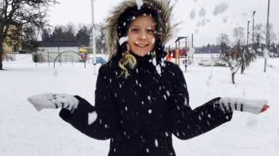 Vinterklätt barn står på snöig skolgård och kastar snö framför sig.