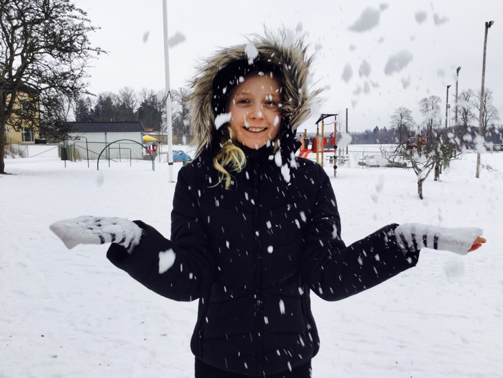 Vinterklätt barn står på snöig skolgård och kastar snö framför sig.