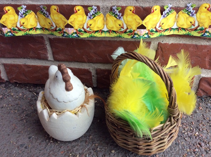 Påskpynt: en korg med gula och gröna fjädrar, en porslinshöna på ett halvt porslinsägg, en pappersdekor med gula kycklingar.