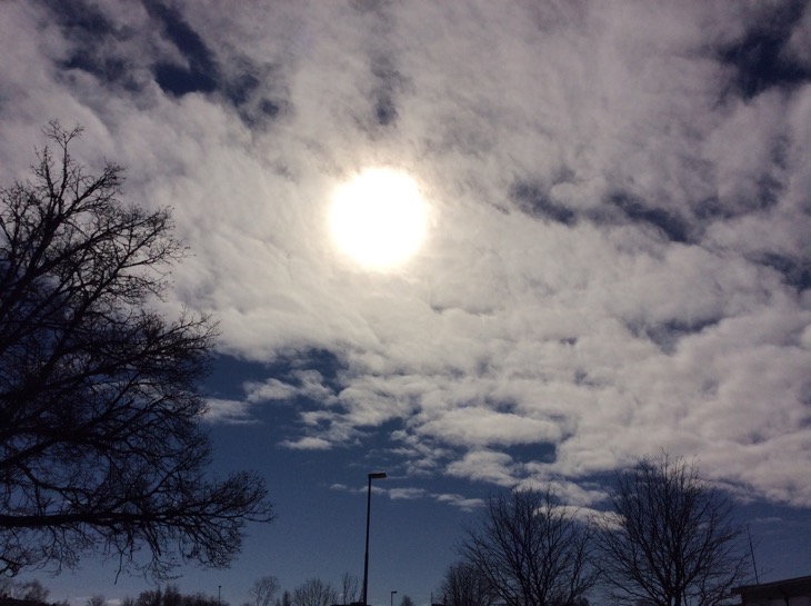 En sol bakom moln omringad av blå himmel. Topparna av träd och en gatlykta.