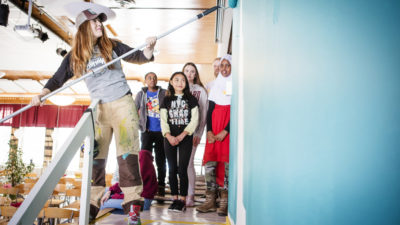 En konstnär målar en vägg turkos. Fyra barn står och tittar på.