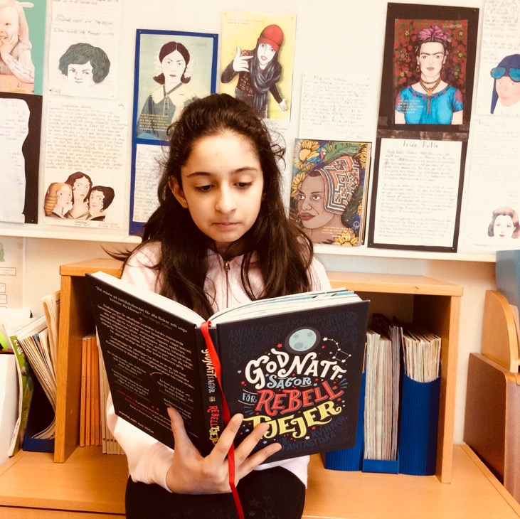 En skolelev sitter och läser i en bok med titeln: Godnattsagor till Rebelltjejer. I bakgrunden är en vägg med uppsatta skolarbeten över historiska kvinnor.