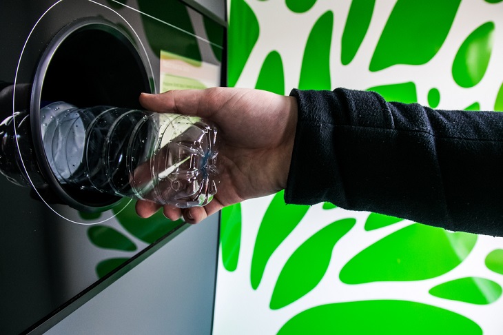 En arm sträcker fram en plastflaska mot en pantmaskin. Flaskan försvinner halvvägs in i ett hål i maskinen.