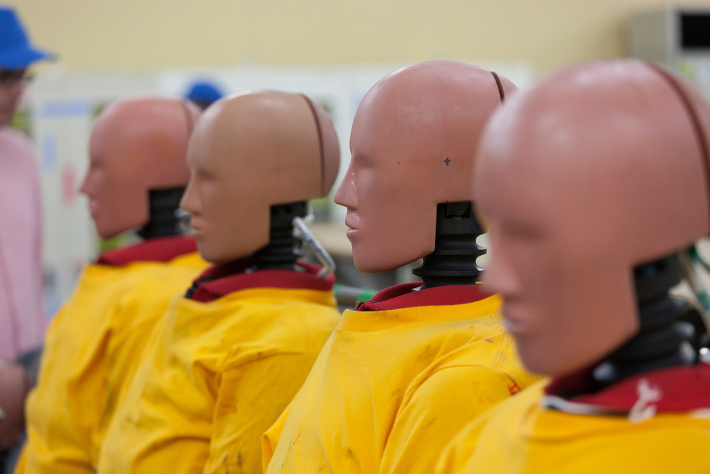 Flera stora dockor sitter i rad. De har huvuden med ansikten som liknar människor, men utan ögon. Nacken är en svart fjäder. De har gula rockar på sig.