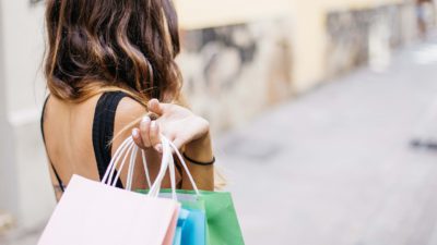 En person med långt hår går nerför en gata. Vi ser bara ryggen. Över axeln har hon slängt några shoppingpåsar.