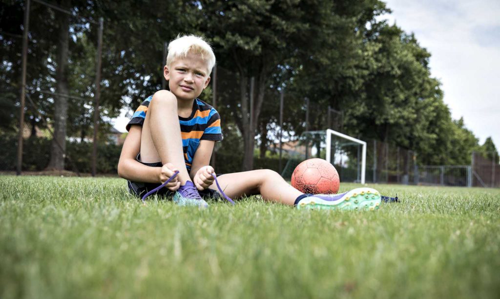 Julle sitter på en gräsmatta. Han knyter sina fotbollskor. Bredvid honom ligger en fotboll. I bakgrunden syns ett fotbollsmål.