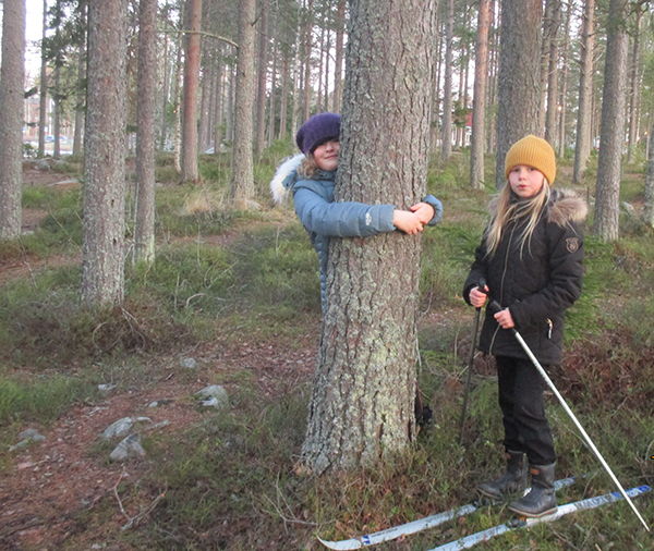 Två elever som står i skogen. Det är barmark. Men den ena flickan kramar ett träd, den andra tjejen har ett par skidor på sig.